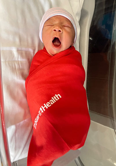 Heart Month Baby swaddled with TriStar Health blanket, yawning.