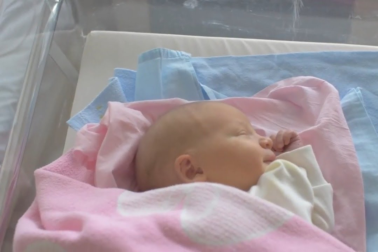 A baby smiles while sleeping in a hospital crib, swaddled in pink and blue blankets.