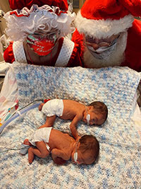 Two infant patients laying on a multicolored quilted baby blanket inside a medical crib are visited by Mrs. Klaus and Santa Klaus who are kneeling over the crib.