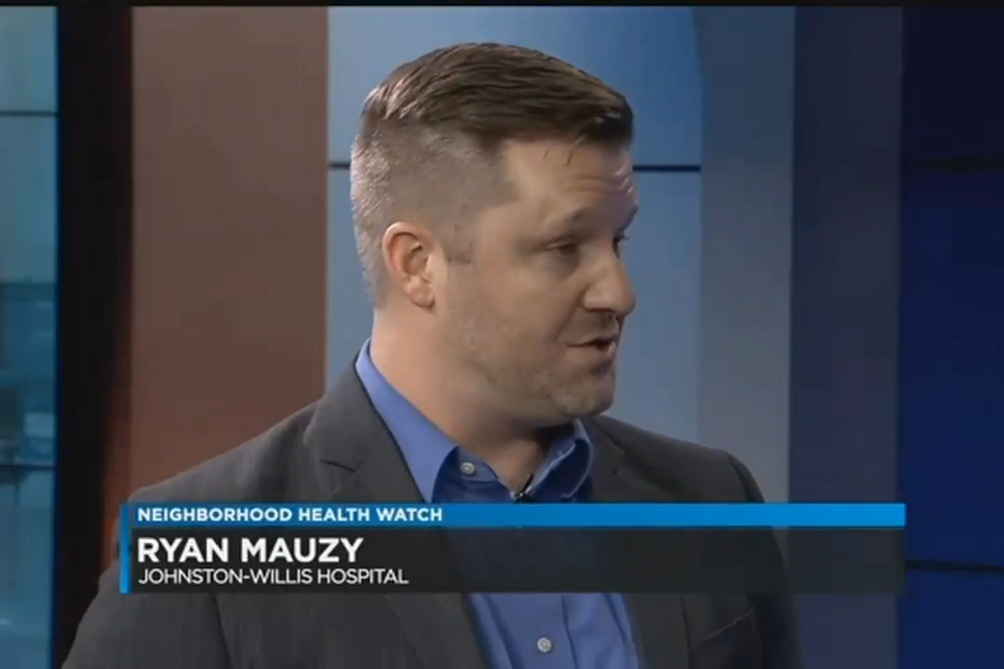 Ryan Mauzy wears a grey blazer and light blue button up shirt while speaking in a television studio as a lower-third graphic reads " Neighborhood Health Watch, Ryan Mauzy, Johnston-Willis Hospital".  