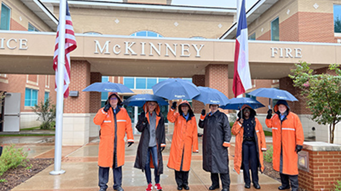 Hospital staff and police officers smile while standing outside of the hospital in the rain, wearing orange and black rain jackets and holding umbrellas.