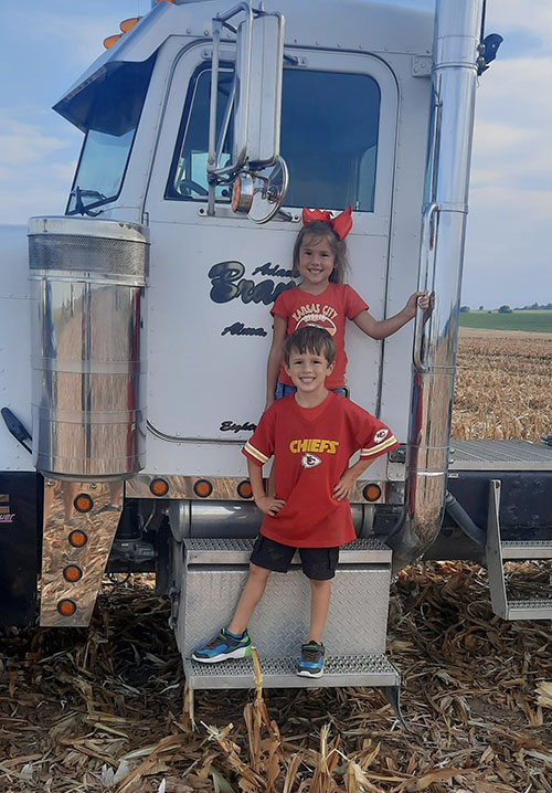 Kellen Brandt's daughter and son smile while wearing Kansas City Chiefs shirts and standing next to a semi hauler truck.