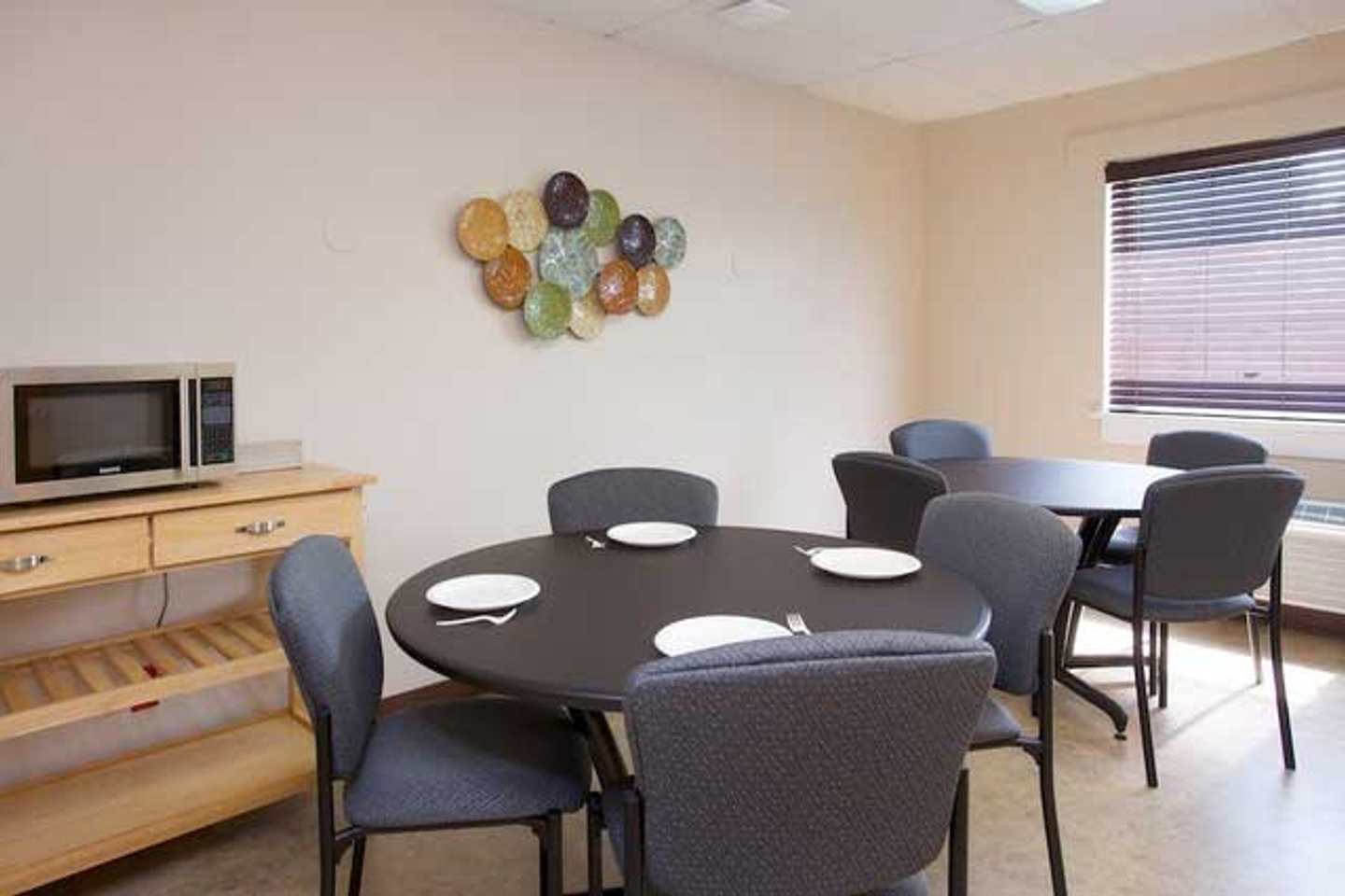 The dining room at Reflections Eating Disorder Treatment Center. Two round tables with four chairs around each of them. One table is set for four people with plates and forks. A table with a microwave is next to them.