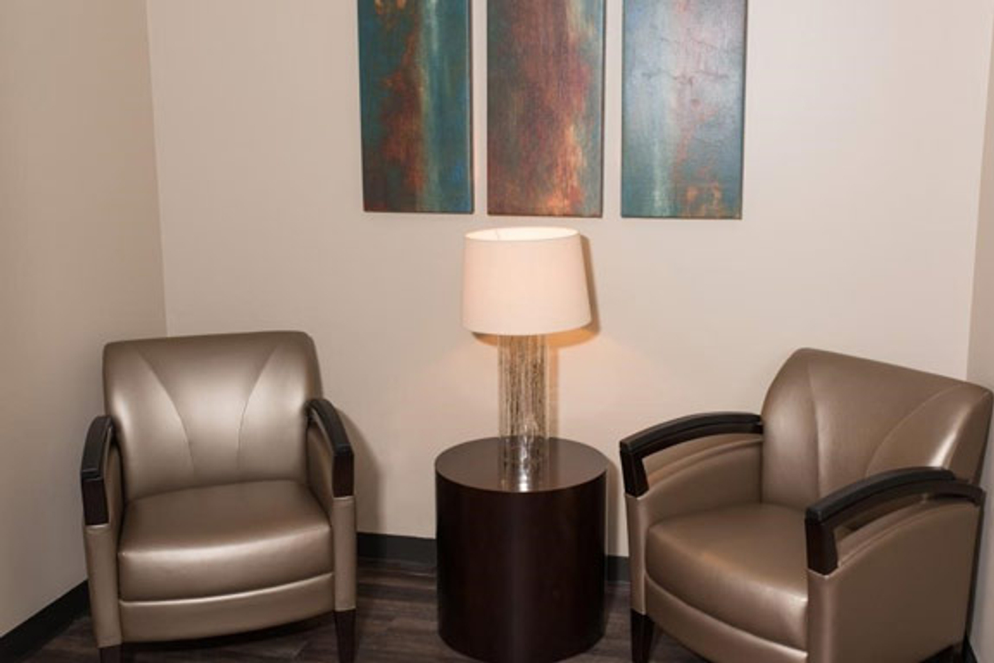 Two brown lounge chairs are seen with an accent table, table lamp between the two chairs. On the wall, three rectangular paintings with abstract bronze and teal colors are hung on the wall behind the chairs.  