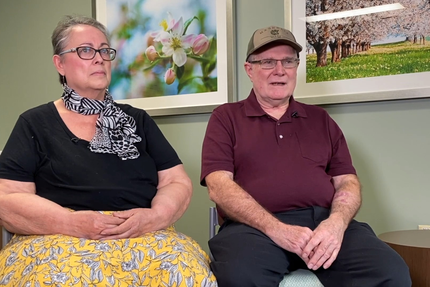 Grace and Dean Hill sit together in front of two large photos of flowering trees, discussing the care their received during their time at Reston Hospital Center.