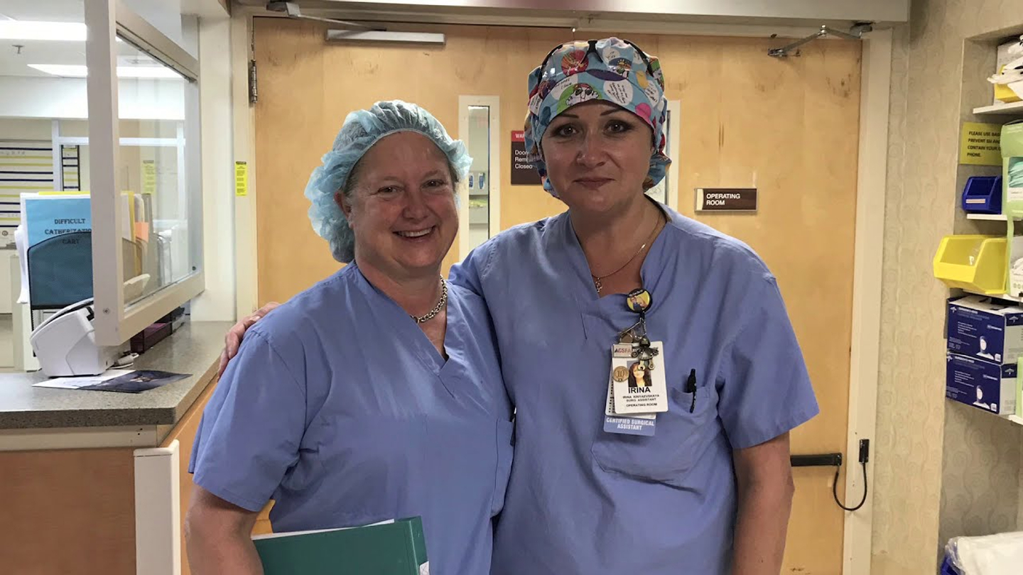 Dr. Cecelia Boardman and a surgical assistant staff member wearing surgery scrubs smile while standing together by a nurse station in the hospital.