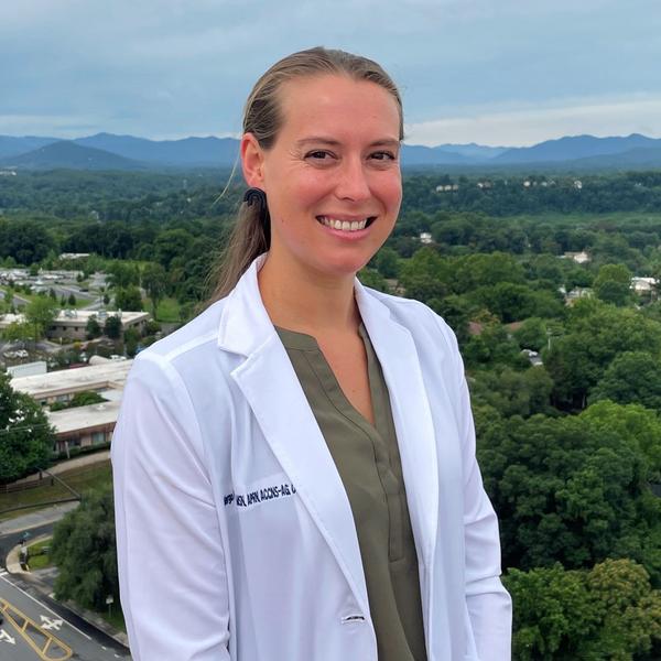 Margo Eatmon smiles while wearing a white lab coat, standing on the roof of the hospital, with the city in the background.