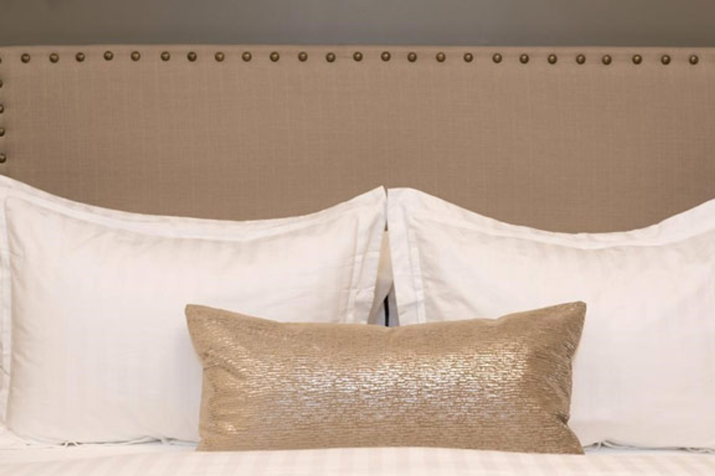 A shimmering beige accent pillow seen from the center of the neatly made bed, rests on two white pillows.