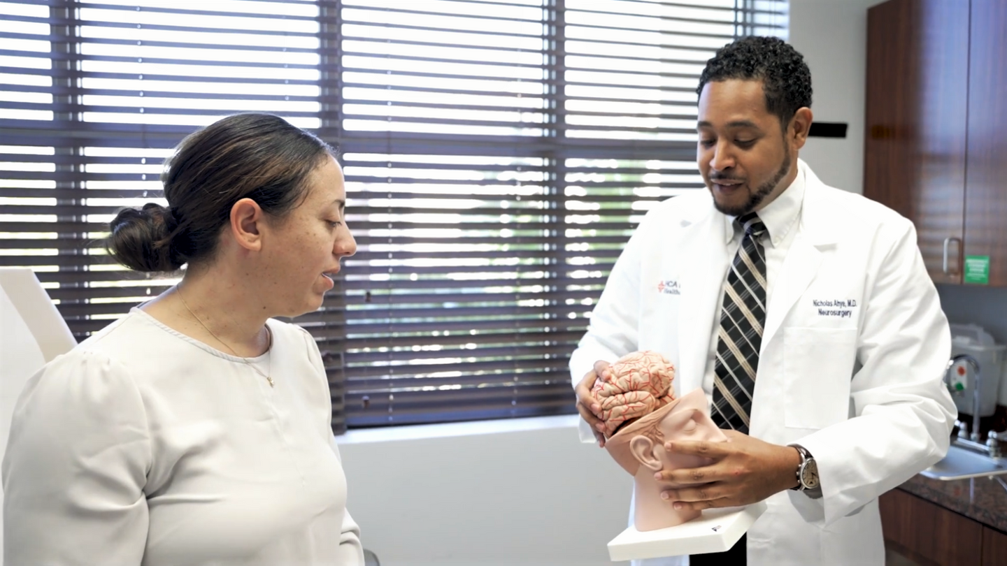 Neurosurgeon Dr. Ahye holding an anatomical model of the human brain, while talking to a female patient in an examination room.