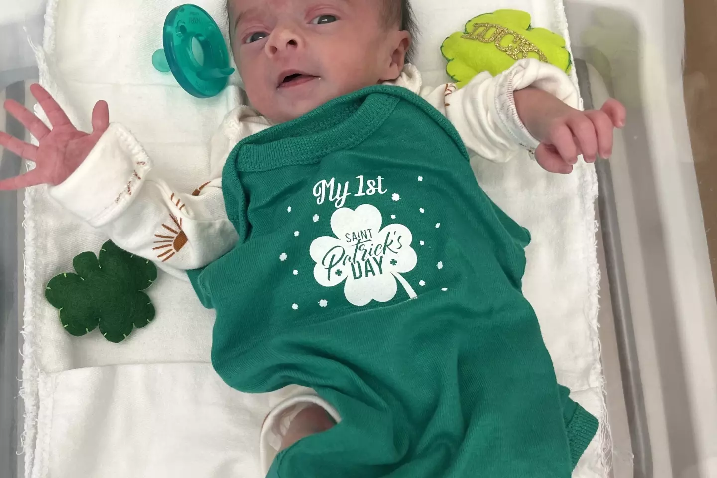 A newborn baby in a green onesies surrounded by plush shamrocks.