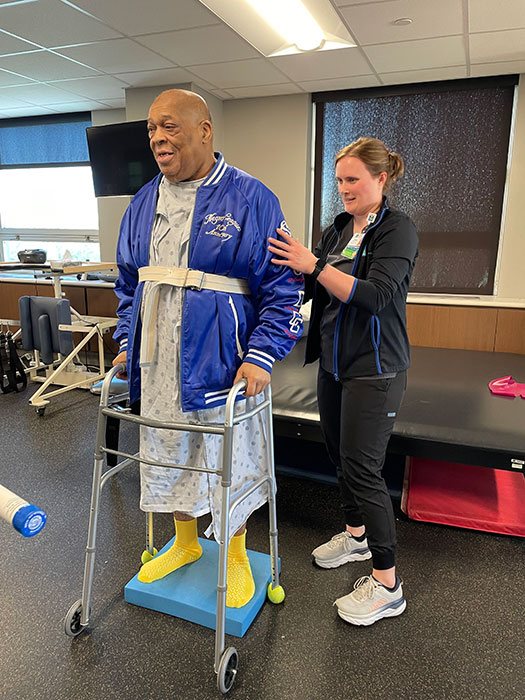 A physical rehabilitation medical professional helps Thomas to stand using a walker.