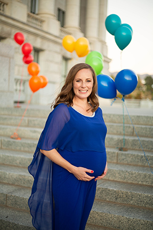 Crystalee Beck holding her pregnant belly outside surrounded by multiple colored balloons.