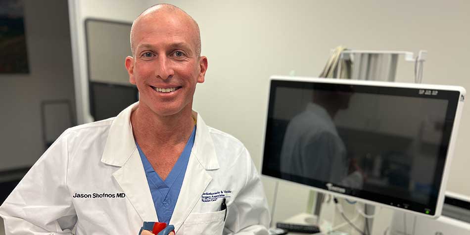 Jason Shofnos, MD, smiles while standing in a patient treatment room with medical equipment in the background.