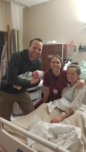 Crystalee Beck sitting up on her hospital bed, with her husband holding their newborn baby boy, and her nurse standing beside her.