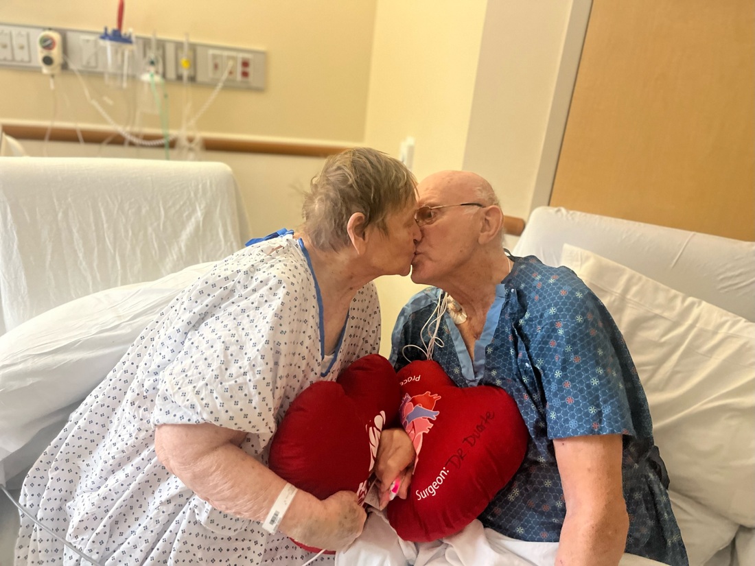 Phil and Margaret Vaske sitting next to each other with heart pillows, kissing in hospital room