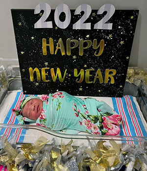 A handmade decorative sign with a "night-sky" background and gold and white lettering that reads, " 2022 Happy New Year" is placed inside a grey and gold tinsel lined decorated hospital bassinet with a newborn baby. The baby is wrapped in a teal colored and floral patterned blanket from head to toe.
