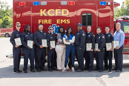 Ramoni smiles while standing with the KCFC Fire EMS team in front of a red ambulance.