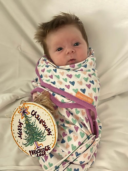A holiday ornament decorated with a holiday tree reads, "Merry Christmas, Parkridge East" next to a infant wrapped in a blanket.