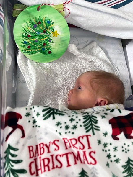 An infant lies in a medical crib underneath a blanket with a holiday design that reads, "Baby's First Christmas."