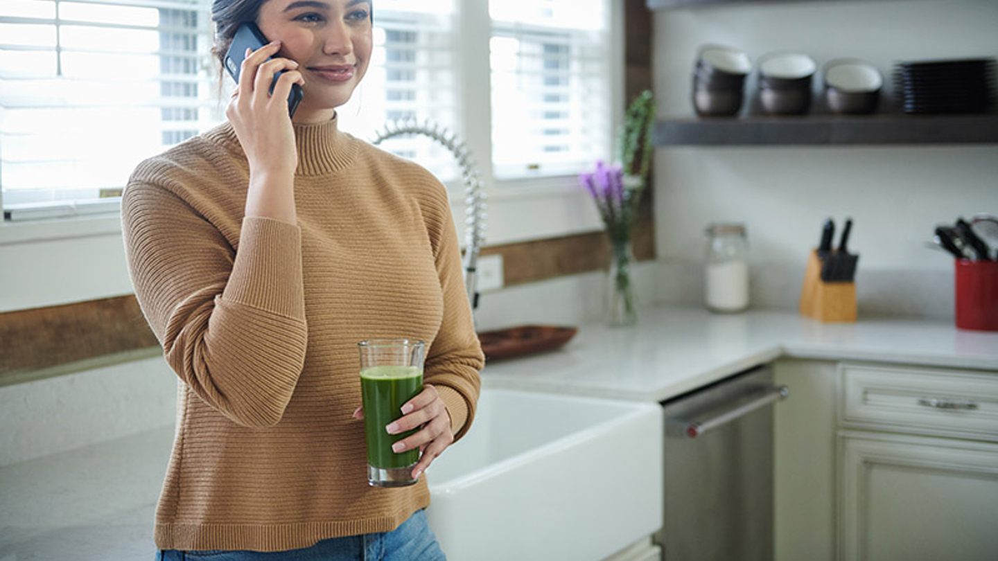 A light-skinned woman holds a cellphone in her right hand while standing in a daytime kitchen. In her left hand, she holds a clear glass with a green juice inside.