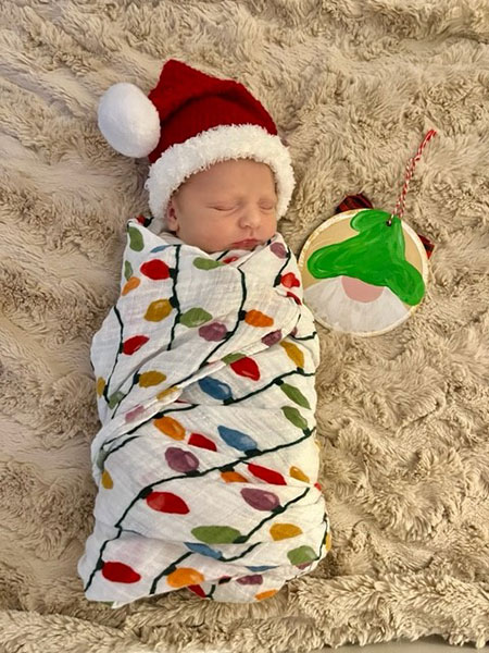 To the left of a holiday ornament, an infant baby is sleeping on a  beige rug wrapped in a holiday light printed blanket wearing a santa cap.