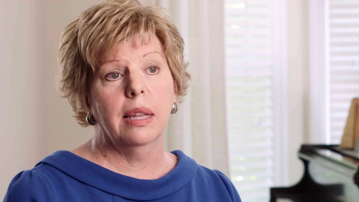 Johnston-Willis patient Michelle Graham, wearing a blue dress with light reddish bob length hair and hoop earrings, is seated in a chair by a black piano illuminated by daylight from partially opened blinds behind Graham