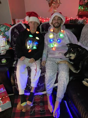 Matt and David Maddux smile while sitting together on the couch during the holidays, both wearing festive hats and necklaces.
