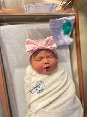 An infant baby, Emi Noel Hicks, is wrapped in a blanket wearing a pink ribbon headband lies in a hospital bassinet.