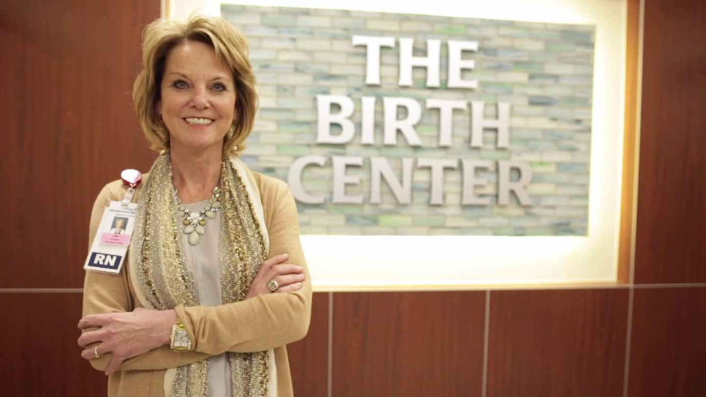 A Registered Nurse stands next to The Birth Center sign with her arms crossed, smiling. 