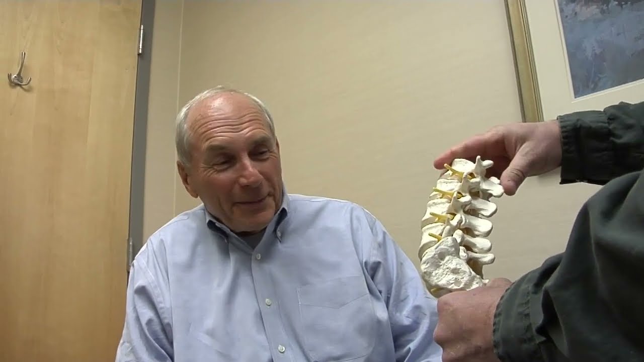 A doctor, out of frame, uses a replica of a spine to explain a surgical procedure to a patient.