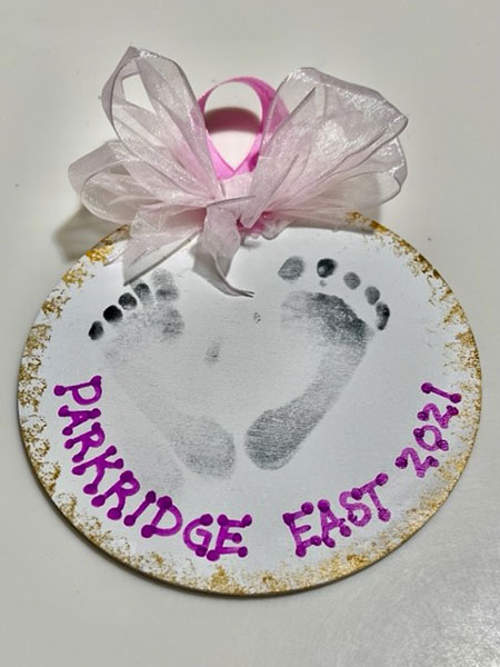 A handmade circular holiday ornament cut out of paper features the black feet marks of an infant with the words, "Parkridge East 2021" written in purple lettering underneath the feet marks. The edges of the ornament are gold dusted, where the top of the ornament also features a pink ribbon and white bow.