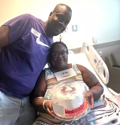 Cynthia Parks holding a cake in hospital bed with significant other at her side