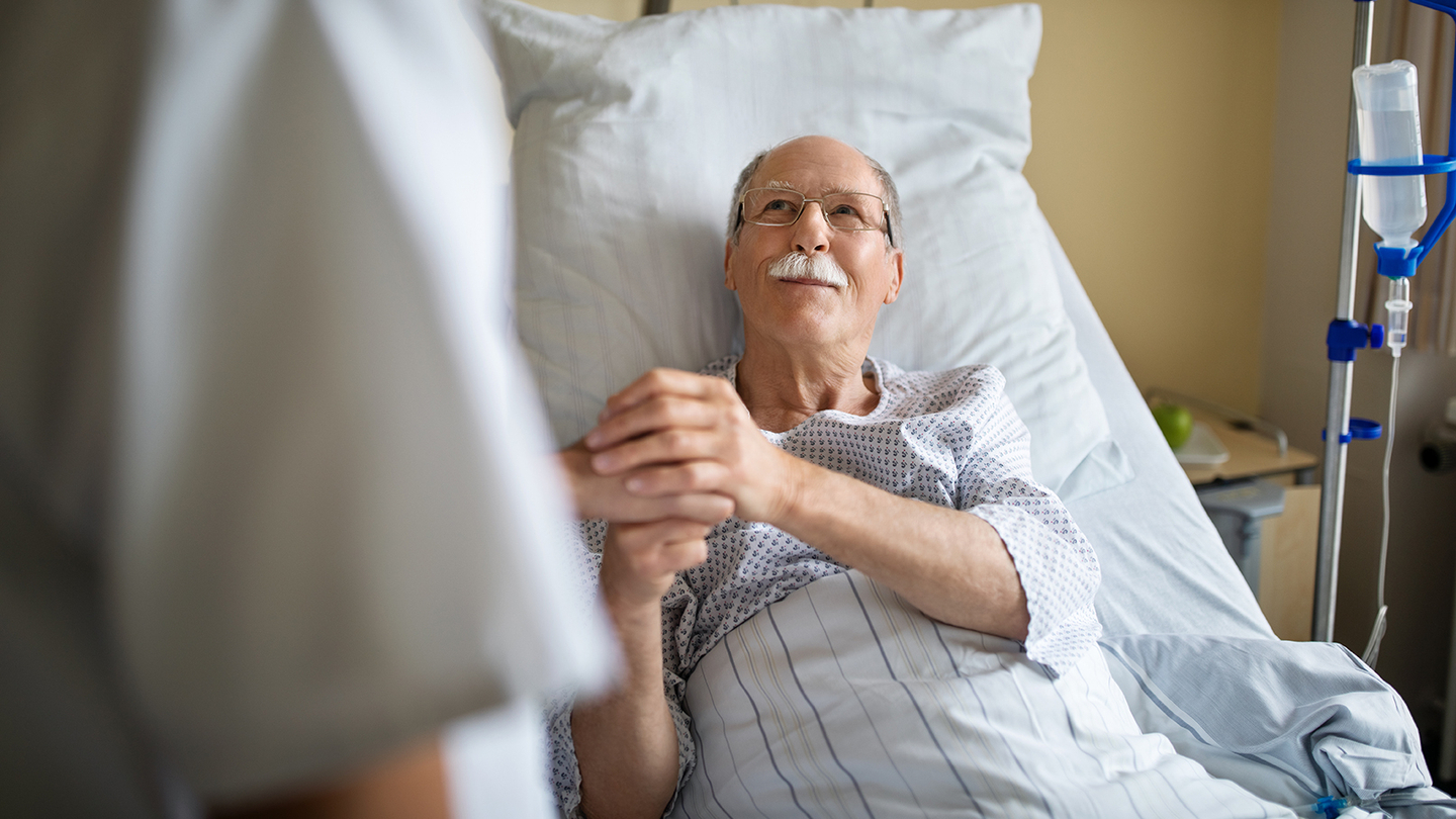 Older man sitting in a hospital bed, holding the hand of a loved one, with a confident loving look in his eyes.