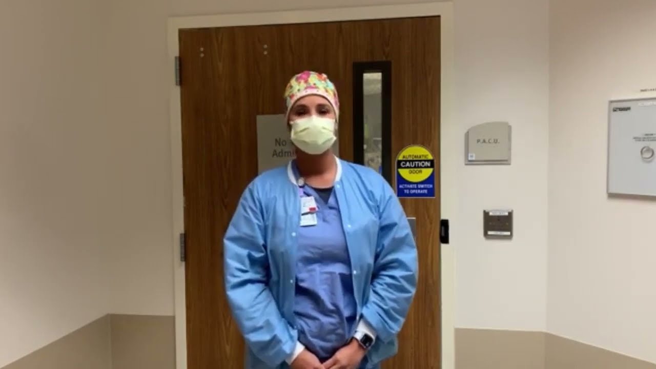 A nurse wearing surgical scrubs and a mask stands in a hallway at Palms West Hospital.