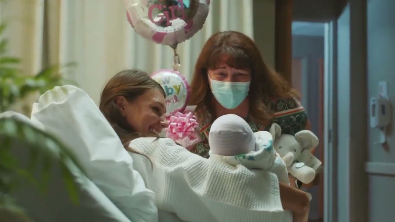 A mother introduces her new baby to a family member who is holding a balloon and gifts.