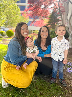 Marilyn Mariani and Mikayla Allred pose with Mikayla's children near trees and grass.