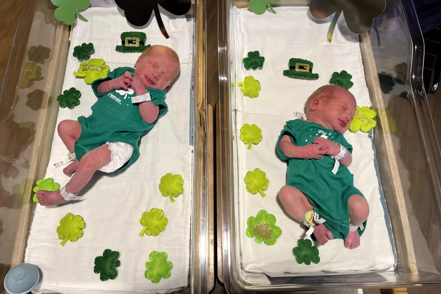 Two newborn babies in green onesies lying in hospital cribs.