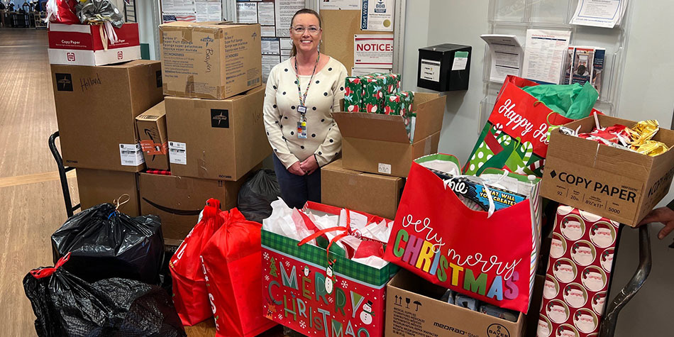 A stack of donated holiday gift bags and donation boxes surround a standing NorthCrest colleague named Michelle, a light-skinned woman with clear glasses, brown hair wearing a holiday sweater and lanyard.